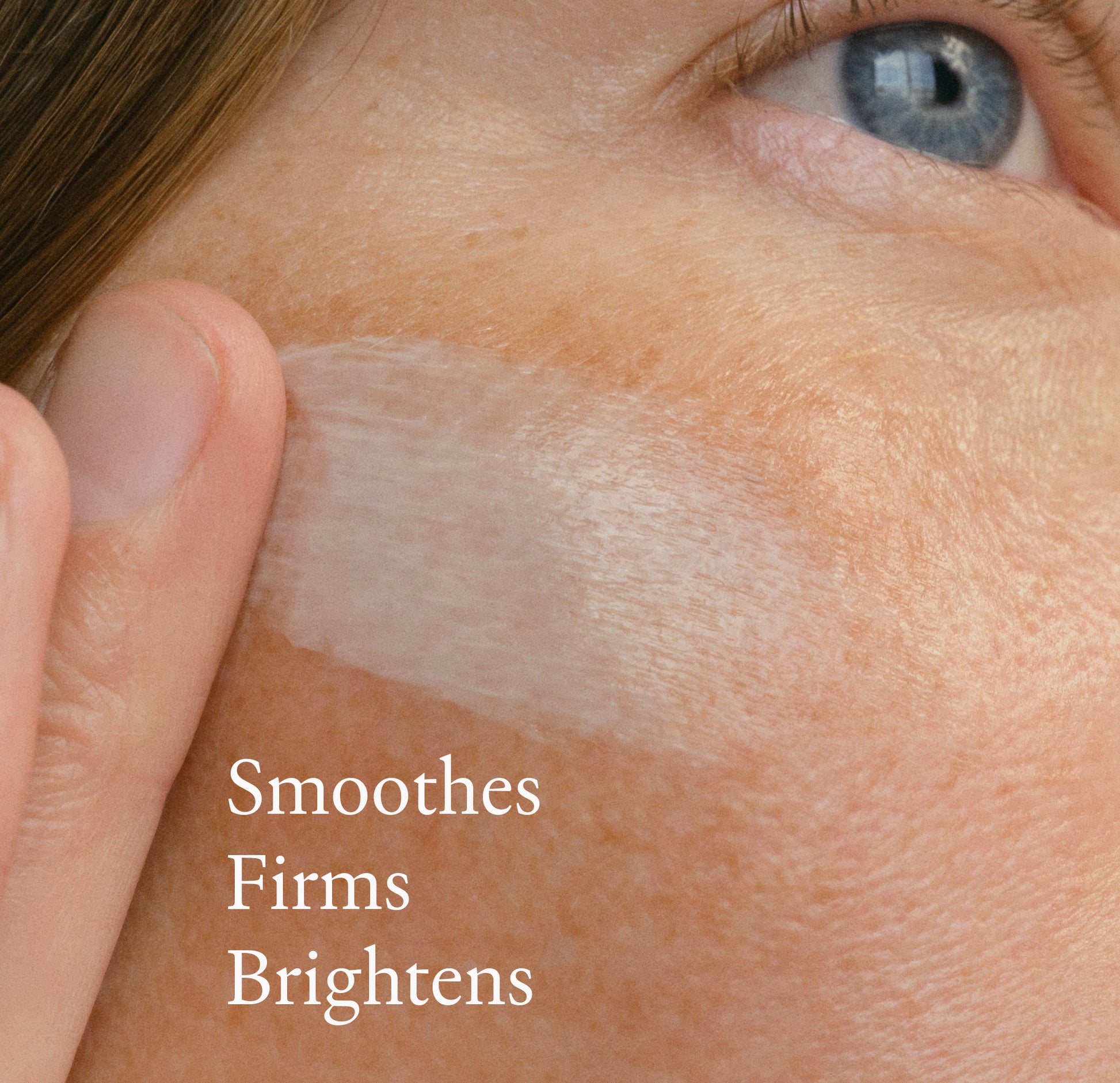 Close-up of a person's cheek with a translucent cream swatch applied across the skin and white text reading 'Smooths Firms Brightens'.