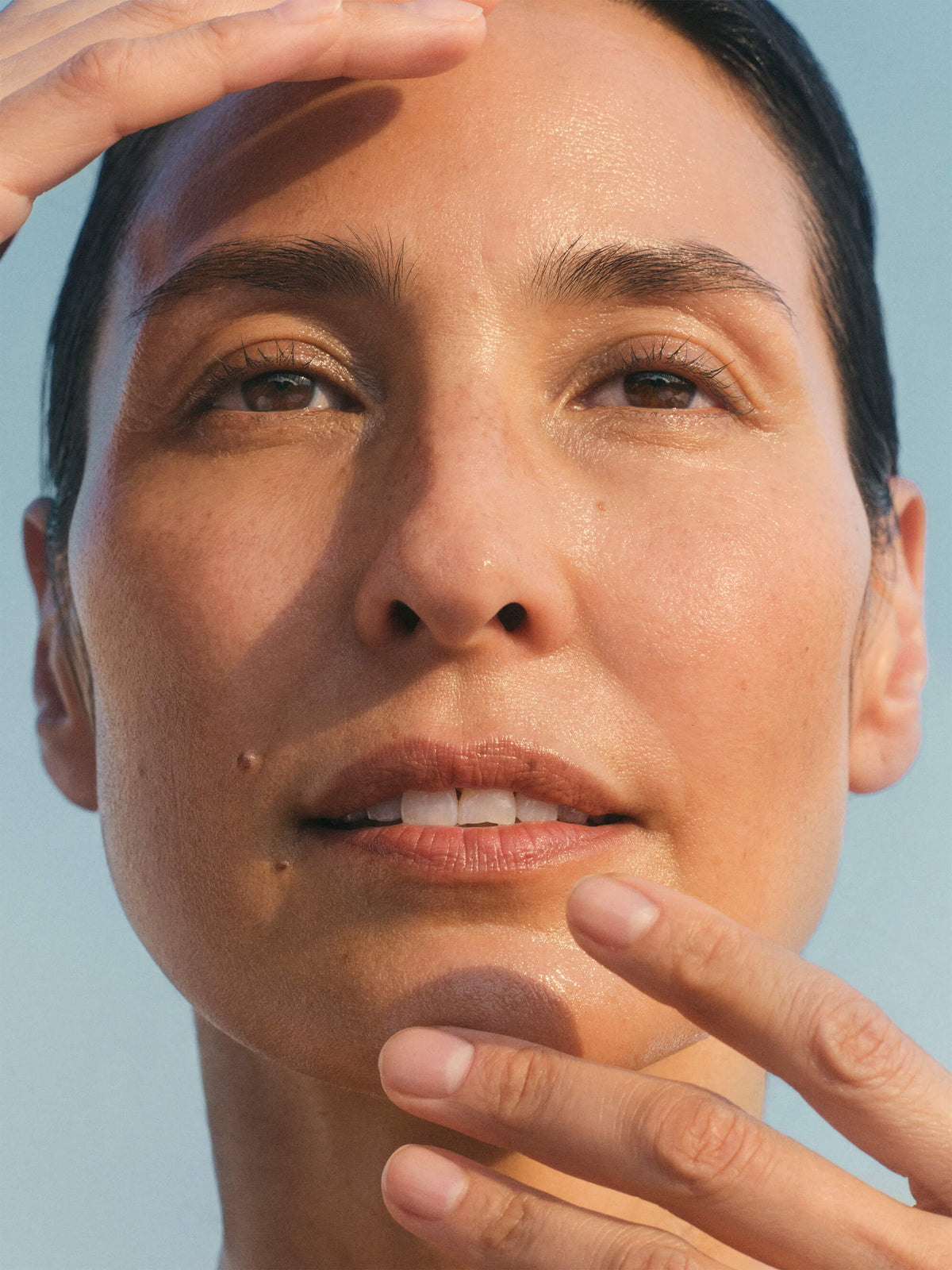 Image of a woman with dark hair pulled back with one hand near forehead and the other lightly touching her chin. 